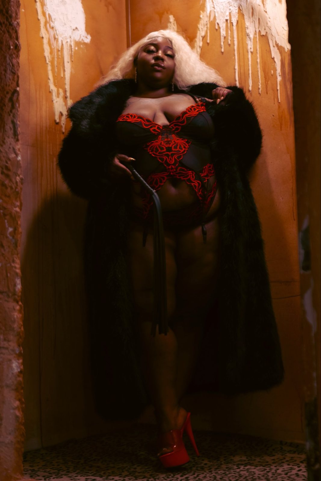 Mistress Sickooo in a black fur coat holding a flogger
